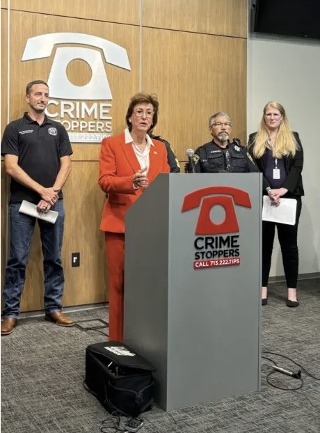 Harris County District Attorney Kim Ogg announced 15 most wanted fugitives for animal cruelty cases. She is wearing a red suit standing behind a podium with the red phone logo, flanked by law enforcement.